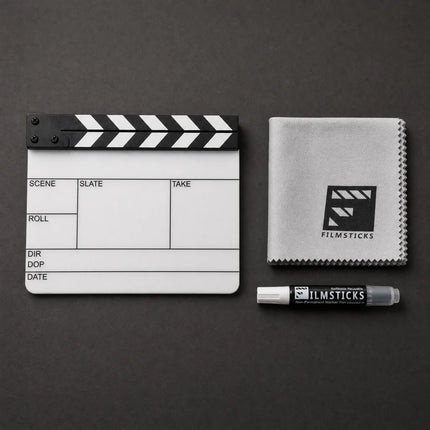 Filmstick kit on a dark gray background, including a black and white clapperboard, a white microfiber cloth with a black Filmsticks logo, and a Filmsticks film stick marker.